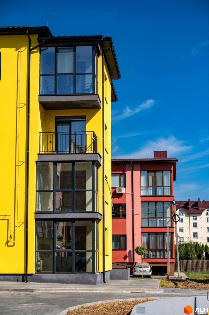 Житловий комплекс Італійський: яскрава жовта новобудова з великими вікнами та балконом на тлі блакитного неба. Поруч червона будівля та прибудинкова територія з деревами та автомобілем.