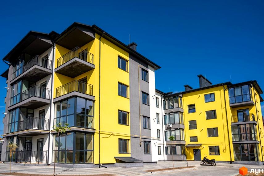 Новобудова ЖК Італійський з яскравою жовтою та сірою фасадною частиною, балконами та панорамними вікнами на тлі блакитного неба. Перед будинком облаштована прибудинкова територія з доріжками та деревами.