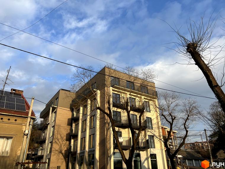 Житловий комплекс Родовід - фасади будівель з балконами та вікнами на тлі хмарного неба. Навколо будівлі видно дерева без листя та елементи міської забудови.