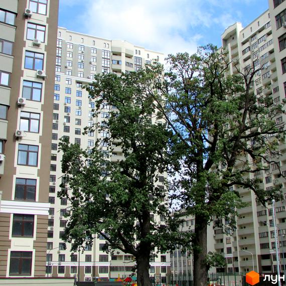 Зовнішній вигляд новобудови Зелений острів-2 з деревами на фоні. Сучасні багатоповерхові будинки з балконами та кондиціонерами.