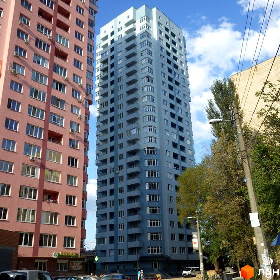 Вигляд на два багатоповерхові будинки: рожевий та світло-блакитний, з численними вікнами та балконами. Житловий комплекс на вул. Феодосійська, 2, оточений деревами та вулицею.