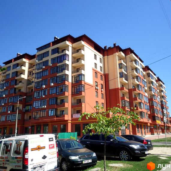 Зображення багатоповерхового будинку Житлового комплексу Академмістечко з червоно-білим фасадом та великими вікнами. Перед будинком припарковані автомобілі та невелика зелена зона з деревами.