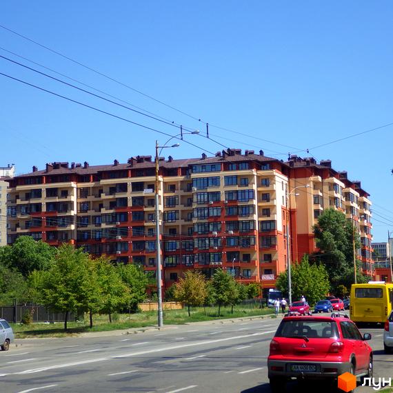 Зображення багатоповерхового будинку Житлового комплексу Академмістечко з фасадом у бежево-помаранчевих тонах. Перед будинком дорога з автомобілями та зелені дерева.