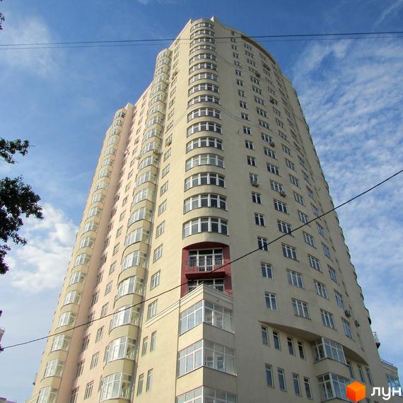 Високий житловий будинок світлого відтінку з вигнутим фасадом та численними вікнами на тлі неба. Житловий комплекс Будинок на Клименка.