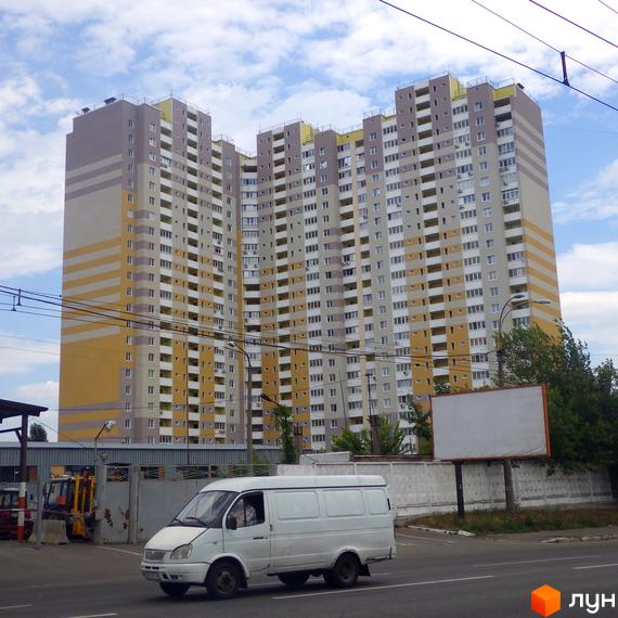 Високоповерховий житловий комплекс на просп. Науки, 55а. Зовнішній вигляд будівлі - сіро-жовті фасади з балконами. На прибудинковій території видно дорогу, білий фургон та будівельну техніку.