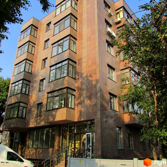 Багатоповерховий будинок з великими вікнами та фасадом з каменю. На передньому плані видно частину прибудинкової території з деревами та білим парканом.