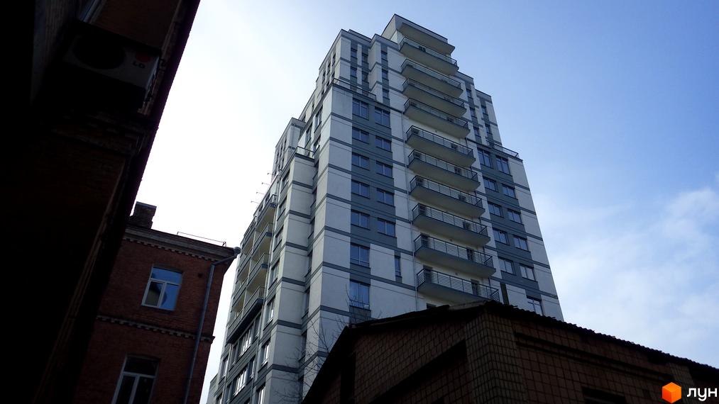 Житловий комплекс Саксаганський: багатоповерховий будинок зі світлим фасадом, темними віконними рамами та балконами. Поруч старі цегляні будівлі.