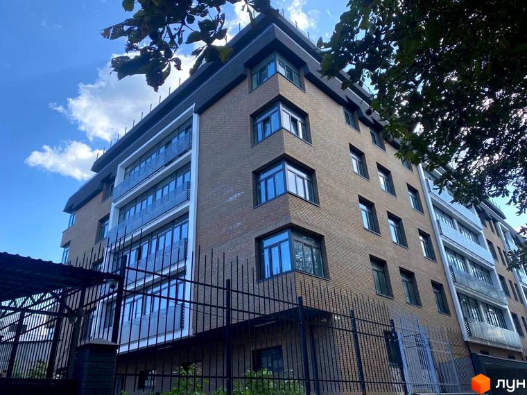 Клубний будинок Donjon з цегляним фасадом та скляними балконами на тлі блакитного неба. На передньому плані видно металевий паркан.