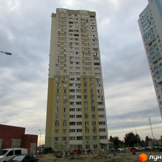 Високий житловий комплекс бежевого та світло-зеленого кольорів на вул. Урлівська, 23в. Поруч прибудинкова територія з автомобілями та дитячим майданчиком.