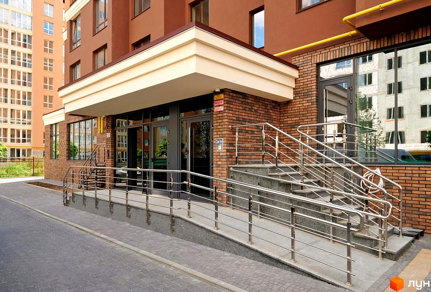 Зовнішній вигляд входу до житлового комплексу Софіївські Липки з пандусом та сходами, огородженими металевими поручнями. Прибудинкова територія вимощена плиткою.