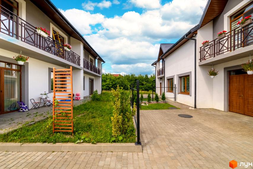 Два двоповерхових будинки з білими фасадами та балконами, що виходять на зелений двір з бруківкою.