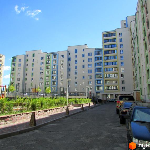 Фасади багатоповерхових будинків Житлового комплексу Акварелі з кольоровими акцентами вікон. На передньому плані дорога, автомобілі та зелені насадження з дитячим майданчиком.