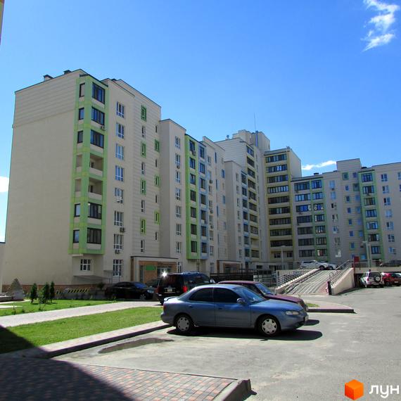 Житловий комплекс Акварелі з багатоповерховими будинками світлого кольору та зеленими вертикальними вставками. На прибудинковій території розташовані парковка, зелений газон та під'їзд до паркінгу.