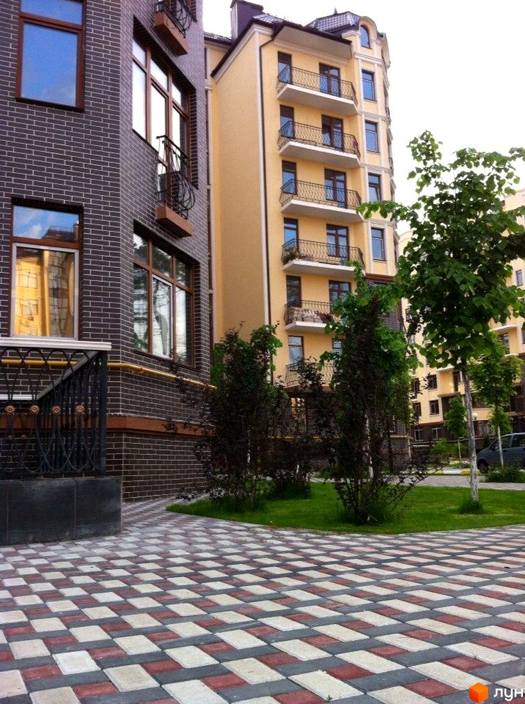 Зовнішній вигляд будівлі ЖК Rich Town з цегляними стінами та балконами. На передньому плані бруківка та зелені насадження.