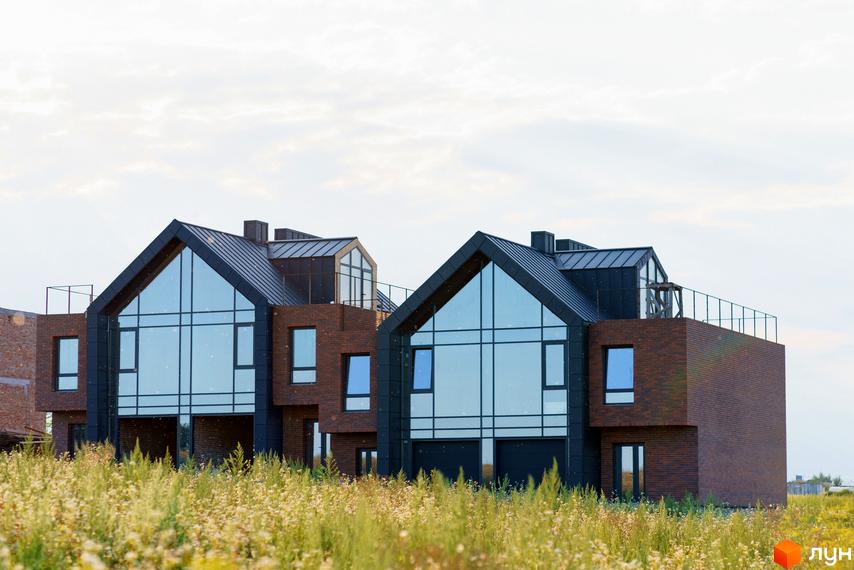 Котеджне містечко Ліщина: два будинки з темними дахами, великими скляними фасадами та цегляними стінами. На передньому плані поле з високою травою.