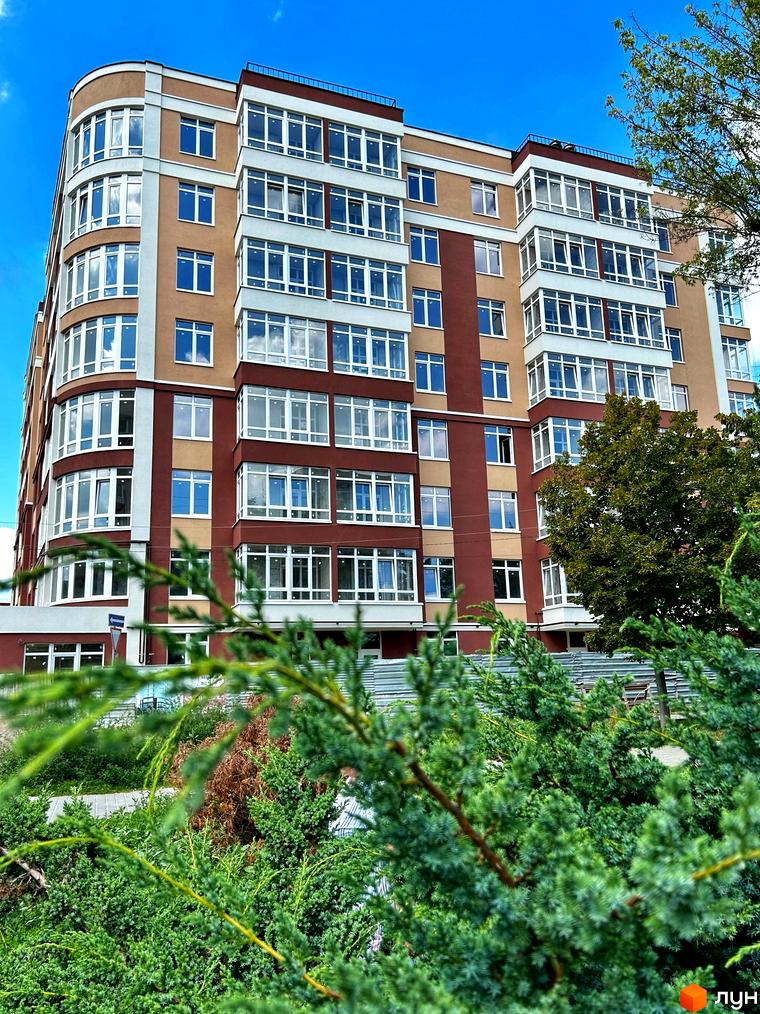 Будинок житлового комплексу Комфорт з коричневими та бежевими фасадами, великою кількістю вікон та балконів. На передньому плані видно зелені кущі.