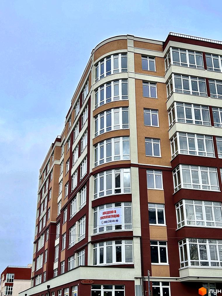 Багатоповерховий будинок з великою кількістю вікон, фасад якого виконано у світлих тонах. На будівлі видно банер з інформацією про введення в експлуатацію.