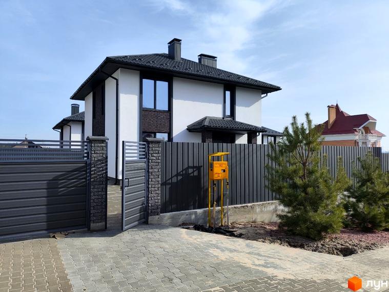 Білий котедж з темним дахом та цегляними елементами у Котеджному містечку Пейзажі Ірпеня. Перед будинком сірий паркан, бруківка та хвойні дерева.