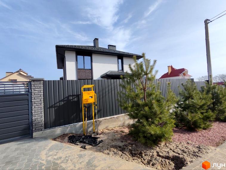 Будинок Котеджного містечка Пейзажі Ірпеня має білі стіни, темний дах та вікна. Прибудинкова територія облаштована парканом, газовим лічильником та молодими соснами.