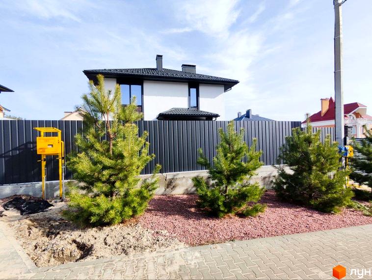 Будинок Котеджного містечка Пейзажі Ірпеня з білим фасадом та темним дахом. Перед ним огорожа, хвойні дерева та ділянка з декоративним щебенем.