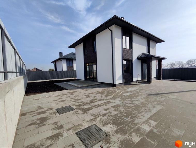 Котеджне містечко Пейзажі Ірпеня: двоповерховий будинок з білими стінами, темним дахом та акцентами з цегли. Перед ним велика вимощена бруківкою територія.