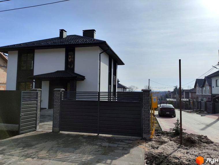 Вигляд Котеджного містечка Пейзажі Ірпеня: двоповерховий будинок з темним дахом та сірою огорожею. Навколо будинку зелена прибудинкова територія.