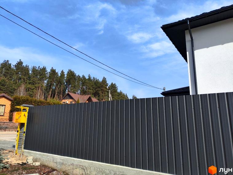 Фасад білого будинку з темним дахом та високий сірий паркан на тлі соснового лісу. Вигляд Котеджного містечка Пейзажі Ірпеня.