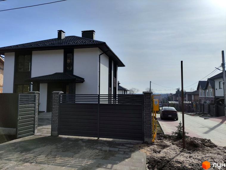 Двоповерховий будинок Котеджного містечка Пейзажі Ірпеня з білими фасадами та темним дахом, перед яким розташовані сірі ворота і вимощений двір.