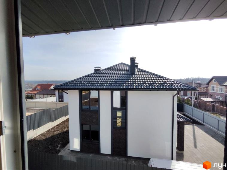 Двоповерховий будинок Котеджного містечка Пейзажі Ірпеня з білими стінами, темними вікнами та дахом. Навколо будинку видно ділянку з огорожею та інші забудови.