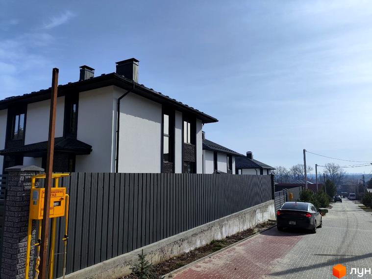 Двоповерховий будинок Котеджного містечка Пейзажі Ірпеня з білими стінами та темним дахом, перед яким розташований паркан та вимощена дорога.