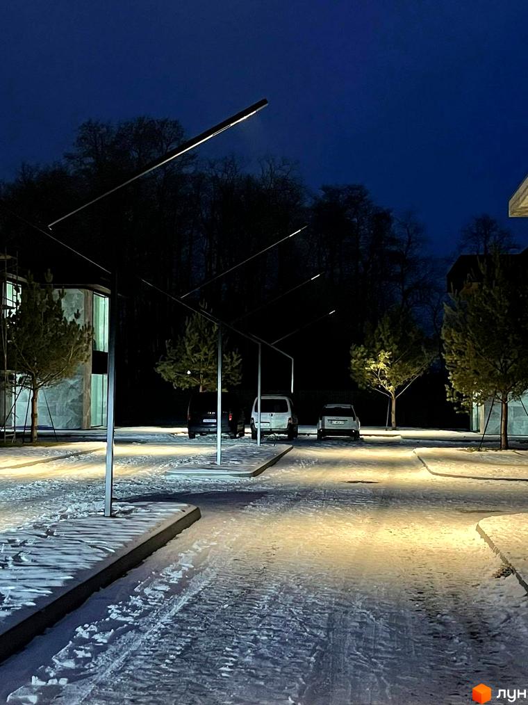 Котеджне містечко MAREA CLUB ROMANKIV вночі, з освітленою територією та деревами, що стоять біля сучасних будівель. Навколо новобудови лежить сніг, а дорога освітлена ліхтарями.