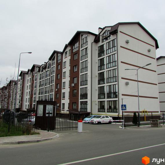 Житловий комплекс Празький квартал. Зовнішній вигляд будинку з коричневими та білими фасадами, великими вікнами та балконами. Прибудинкова територія з парканом, шлагбаумом та автомобілями.