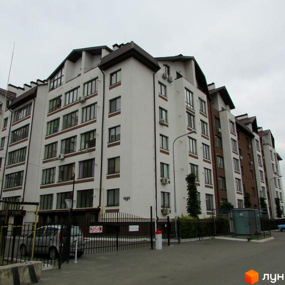 Зовнішній вигляд новобудови ЖК Празький квартал з білим фасадом та коричневими елементами. На прибудинковій території видно паркан та автомобілі.