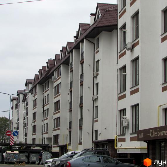 Зовнішній вигляд новобудови ЖК Празький квартал з білими фасадами та коричневими елементами. Поруч видно прибудинкову територію з автомобілями та сміттєвими баками.