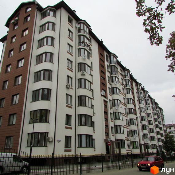 Зовнішній вигляд новобудови ЖК Празький квартал з коричневими та білими фасадами. Навколо будинку розташована огорожа та дорога з автомобілями.