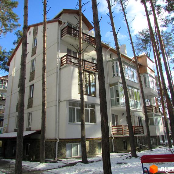 Зовнішній вигляд житлового комплексу Островський: біла будівля з балконами серед високих дерев. Прибудинкова територія вкрита снігом.