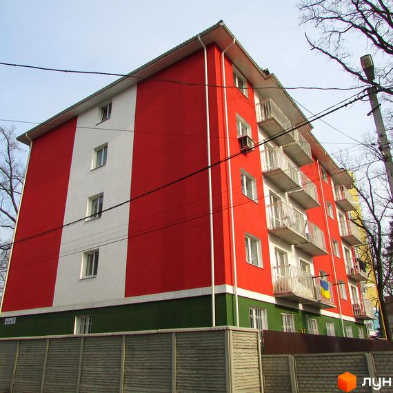 Житловий комплекс Щасливий має яскравий фасад у червоно-білу смугу із зеленим цоколем та білими балконами. Навколо будівлі видно паркан та дерева.