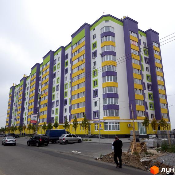 Житловий комплекс Щасливий на Яблуневій – яскрава новобудова з жовто-фіолетовими фасадами. Навколо будинку облаштована прибудинкова територія з деревами та місцями для паркування.