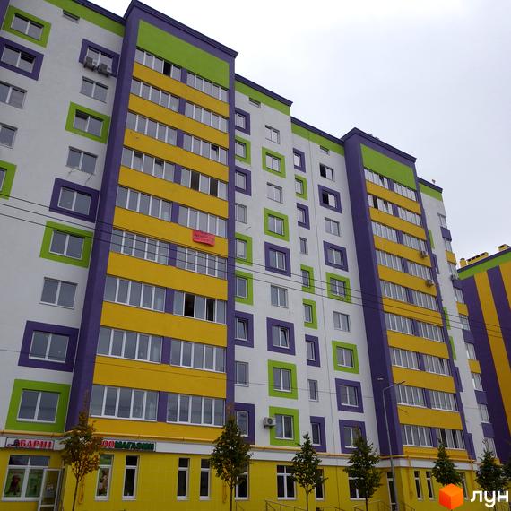 Житловий комплекс Щасливий на Яблуневій, багатоповерховий будинок з яскравим фасадом у біло-зелено-жовто-фіолетових кольорах. Навколо будинку зелені насадження та дерева.