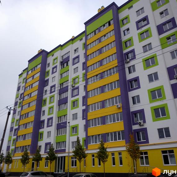 Яскравий житловий комплекс Щасливий на Яблуневій з фасадами жовтого, зеленого та фіолетового кольорів. Перед будинком висаджені молоді дерева.