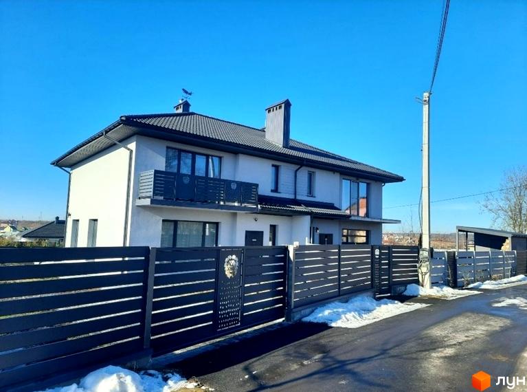 Фасад будинку Котеджного містечка ЗУБРА ETALON зі світлими стінами та темним дахом. Територія огороджена темним парканом з воротами, прикрашеними левом.