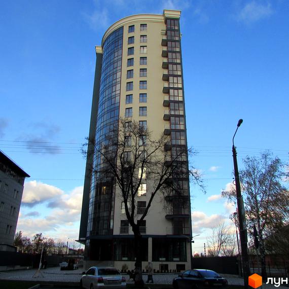 Житловий комплекс Фамільний - висотна новобудова з панорамним склінням та балконами. Перед будинком розташована прибудинкова територія з деревами та автомобілями.