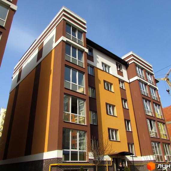 Фасад житлового комплексу Затишний з коричневими та жовтими елементами, великими вікнами та білими декоративними смугами. Будинок має багатоповерхову структуру під блакитним небом.