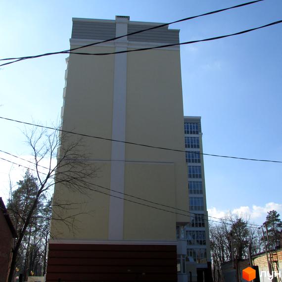 Новобудова Житловий комплекс Капітал: світло-жовтий фасад з білою вертикальною смугою, сірим дахом та темною основою. Навколо дерева.