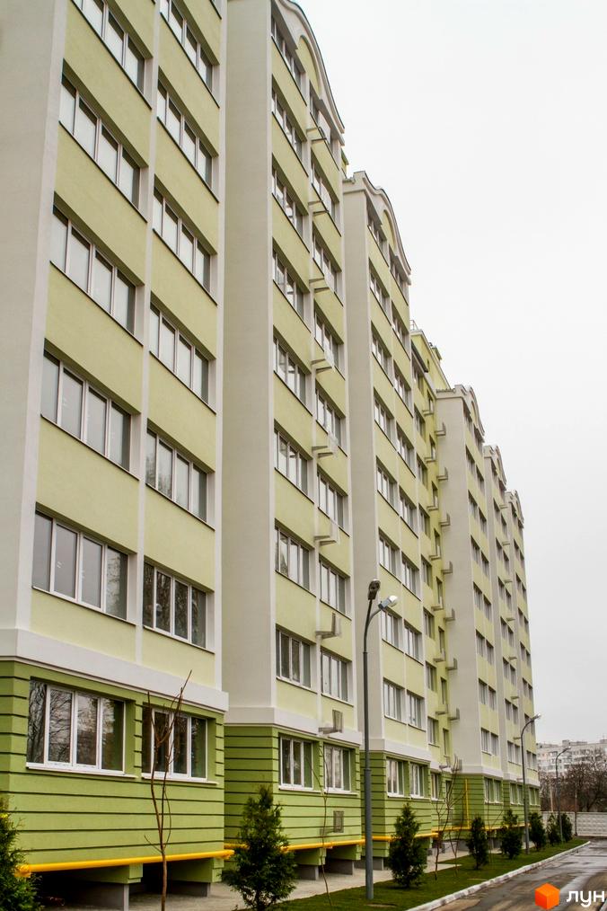 Багатоповерховий будинок житлового комплексу Первоцвіт з великою кількістю вікон. Прибудинкова територія з зеленими насадженнями та ліхтарями.