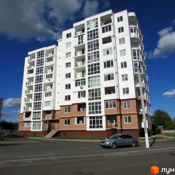 Новобудова ЖК Вишиванка. Багатоповерховий будинок з білим фасадом, великими вікнами та балконами на тлі блакитного неба з хмарами. Перед будинком дорога та припаркований автомобіль.