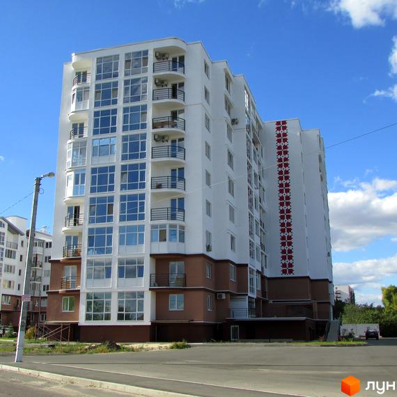 Новобудова ЖК Вишиванка з білою фасадною частиною та коричневим цоколем, балконами та великими вікнами. Навколо будинку облаштована прибудинкова територія з дорогою та зеленими насадженнями.