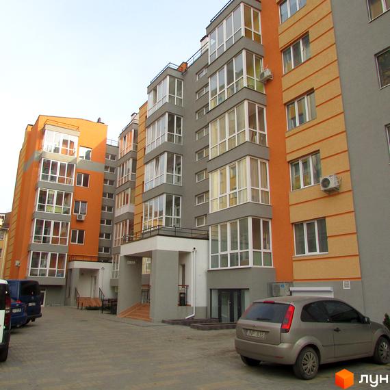 Зовнішній вигляд новобудови ЖК Status з помаранчевими та сірими фасадами, великими вікнами та балконами. Прибудинкова територія з паркуванням та автомобілями.