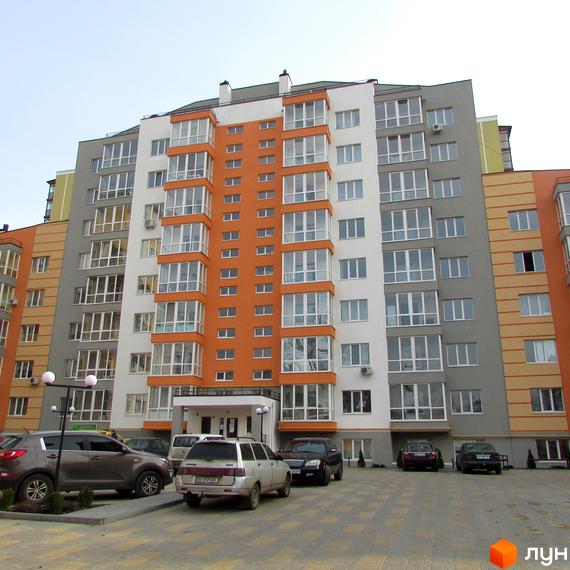 Житловий комплекс Status – це багатоповерховий будинок з яскравим фасадом та балконами, розташований на фоні ясного неба. Перед будинком знаходиться парковка з автомобілями.