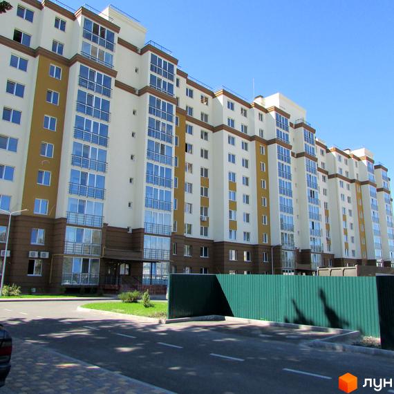 Житловий комплекс Променада: багатоповерховий будинок біло-жовтого кольору з коричневим цоколем та скляними балконами. Перед будинком дорога та зелений паркан.